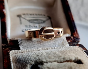 May include: A gold-toned ring with a belt buckle design. The buckle is detailed with a raised center and two small holes.