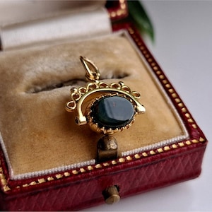 May include: A gold pendant featuring a dark green oval gemstone, set in an ornate frame. The pendant is presented in a red jewellery box with gold trim and a beige interior.