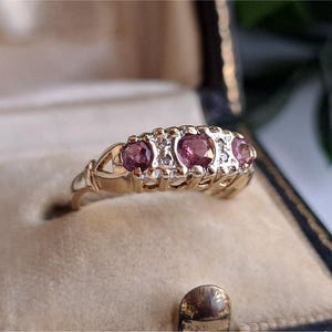 May include: A gold ring featuring five pink gemstones and small, clear accent stones. The ring is set in a gold band with decorative filigree. It is presented in a vintage jewellery box with a beige interior.