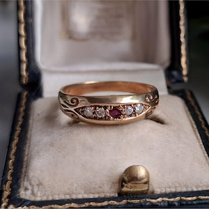 May include: A gold band ring featuring a central red gemstone flanked by two small clear stones on each side. The metal surface displays engraved scrollwork patterns on both ends of the stone setting, creating a detailed and ornate appearance.