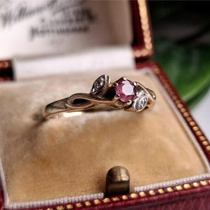 May include: A gold ring featuring a central ruby and two diamond accents. The ring has a vintage design, displayed in a red jewellery box with gold trim and a tan interior. The ring is a piece of jewellery.
