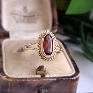 Op de afbeelding: Een gouden ring met een ovaal geslepen dieprode granaat edelsteen in een zetting. De steen is omringd door een decoratieve gedraaide, touwachtige metalen rand. De ringband is dun en splitst zich in twee delen bij de centrale zetting.