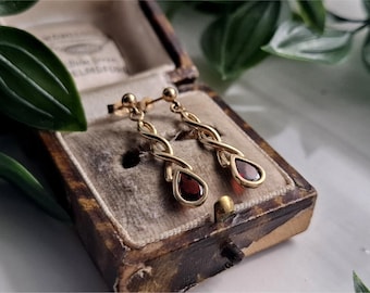Könnte beinhalten: Ein Paar goldfarbene Ohrhänger mit einem gedrehten Design, die jeweils einen tropfenförmigen, tiefroten Edelstein aufweisen. Die Ohrringe werden in einer Vintage-Schmuckschatulle mit Samtfutter präsentiert.