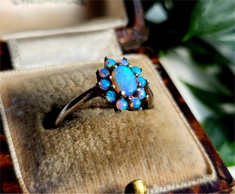 May include: A silver ring featuring a central oval opal surrounded by smaller opals. The opals display a range of blue and pink hues. The ring is set in a vintage-style setting, showcasing a floral design.