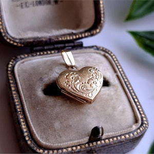 May include: A gold heart-shaped locket with floral detailing, resting in a vintage jewelry box. The locket is suspended from a small loop and features an ornate design. The box has a velvet interior and a decorative, patterned exterior.