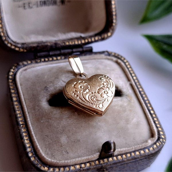 Könnte beinhalten: Ein goldfarbenes herzförmiges Medaillon mit floralen Details, das in einer Vintage-Schmuckschatulle liegt. Das Medaillon ist an einer kleinen Schlaufe aufgehängt und weist ein verziertes Design auf. Die Schatulle hat ein Samtinterieur und ein dekoratives Äußeres.