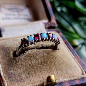 May include: A silver ring with alternating pink and blue gemstones. The ring is in a small, brown velvet box.