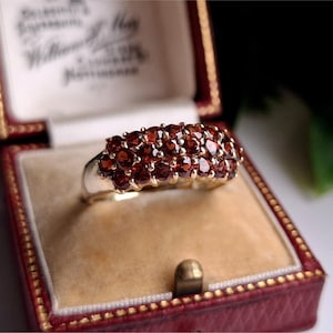 Puede incluir: Un anillo de oro con un racimo de pequeñas gemas redondas de color rojo intenso. El anillo se exhibe dentro de un joyero rojo vintage con un interior color crema.