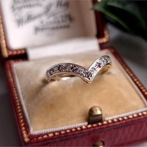 May include: A gold-toned ring with a V-shaped design, set with small red and clear gemstones. The ring is displayed inside a red jewelry box with a cream-colored interior.