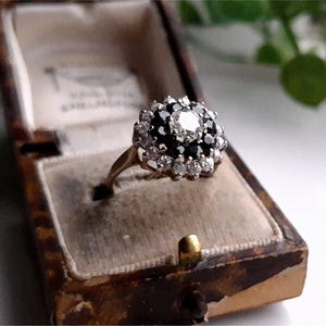 May include: A gold ring with a large, round, white gemstone surrounded by smaller black gemstones. The ring is in a brown velvet ring box.