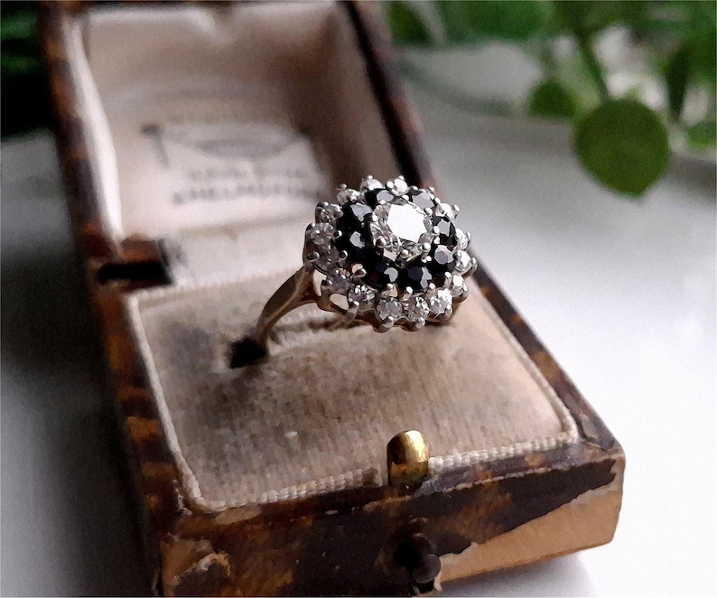 May include: A gold ring with a large, round, white gemstone surrounded by smaller black gemstones. The ring is in a brown velvet ring box.