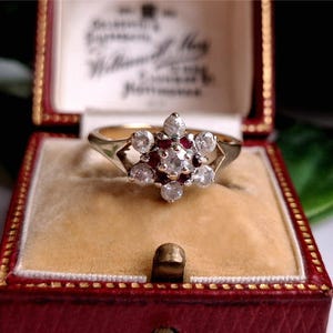 May include: A gold ring with a floral design, featuring a central cluster of diamonds and rubies. The ring is displayed inside a red jewelry box with a cream-colored interior, showcasing its intricate details and vintage style.