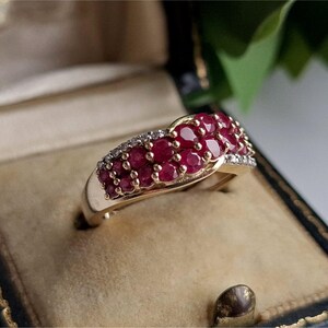 May include: A gold ring with a cluster of small, round, deep red gemstones, possibly rubies, set in a curved design. The ring also has a row of smaller, clear gemstones, likely diamonds, adding sparkle. The ring is displayed in a vintage jewellery box.