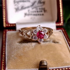 May include: A gold ring with a flower-shaped design, featuring a central red gemstone surrounded by white gemstones. The ring is displayed in a red velvet-lined jewelry box.