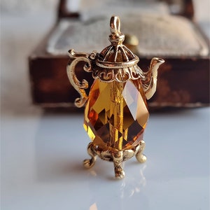 May include: A gold-tone teapot charm featuring a clear amber crystal body and intricate detailing. The charm is designed with a spout, handle, and a decorative base.