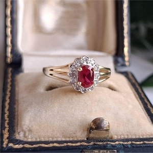 May include: A gold ring with a large oval ruby center stone, surrounded by a halo of small diamonds. The ring is set in a vintage-style setting and is displayed in a black and gold jewelry box. The ring's band has a split design.