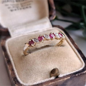 May include: A gold band ring featuring an alternating pattern of three round red rubies and three marquise-shaped clear white diamonds set in a row across the top of the band, presented in a vintage-style brown velvet-lined jewellery box.