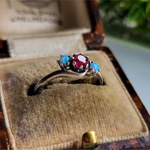 Peut inclure: Une bague en argent ornée d'une pierre rouge centrale et de deux opales bleues. La bague est présentée dans un écrin à bijoux marron vintage avec un intérieur en velours, mettant en valeur son design et le contraste des couleurs.