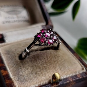 May include: A silver-toned ring featuring a cluster of seven round, deep red faceted stones arranged in a floral pattern. The metal band has a slender, tapered design that connects to the central stone setting, presented inside a vintage box.
