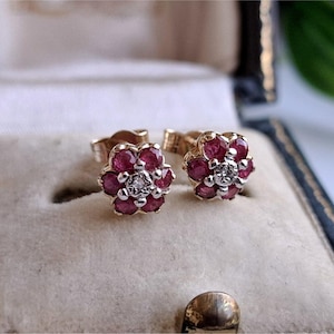 May include: A pair of stud earrings featuring a floral design with six round red rubies surrounding a central clear diamond. The stones are set in a metallic frame with a post backing, creating a cluster shape that resembles a small flower blossom.