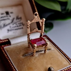 Può includere: Un ciondolo a forma di sedia in miniatura color oro con un sedile rettangolare in pietra preziosa rossa. La sedia ha gambe dettagliate, uno schienale e un anello per una catena. Il ciondolo è esposto in un portagioie rosso e beige.
