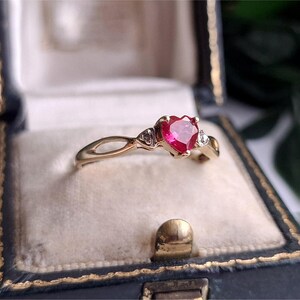 May include: A gold ring featuring a central heart-shaped red gemstone flanked by two small clear stones on each side. The band has a split design near the setting, and the ring is presented inside a dark blue velvet-lined jewellery box.