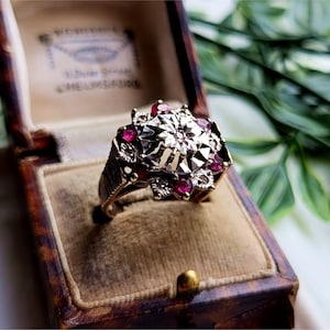 Könnte beinhalten: Ein Vintage-Goldring mit detailliertem floralem Design, besetzt mit kleinen roten Edelsteinen. Der Ring wird in einer braunen, samtgefütterten Schmuckschatulle präsentiert, die seine komplizierte Handwerkskunst und seinen klassischen Stil zeigt. Das Band des Rings hat ein strukturiertes Design.