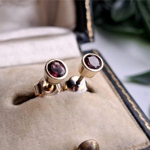 Può includere: Un paio di orecchini a lobo con pietre preziose rotonde sfaccettate di colore rosso scuro, incastonate in castoni di metallo color oro lucido. Gli orecchini hanno perni dritti e sono fissati con chiusure a farfalla in metallo abbinate.