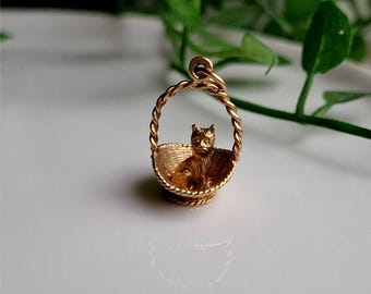 Puede incluir: Un dije dorado que representa un gato sentado en una cesta tejida. La cesta tiene un asa trenzada y un patrón tejido detallado. El gato también es dorado y tiene una cara detallada.
