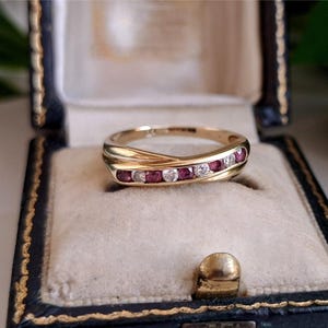 May include: A gold band ring featuring a crossover design with a channel setting. The ring is inlaid with a row of alternating round-cut red rubies and clear white diamonds, creating a continuous sparkling pattern across the centre of the band.