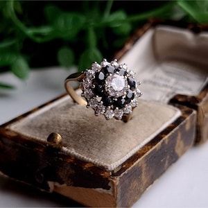 May include: A gold ring with a large center stone surrounded by smaller black and white stones. The ring is in a small, brown velvet box.