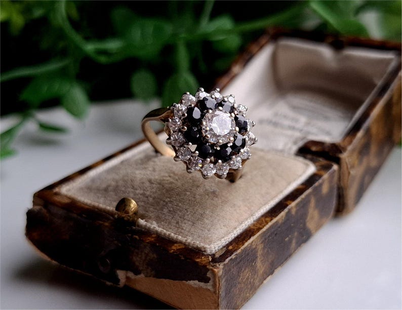 May include: A gold ring with a large center stone surrounded by smaller black and white stones. The ring is in a small, brown velvet box.