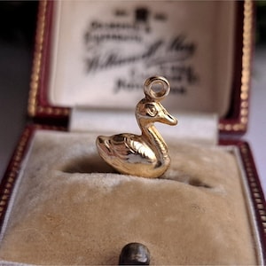 May include: A gold-colored duck charm with intricate detailing, displayed inside a vintage red jewelry box with a cream-colored interior. The charm has a loop for attaching to a necklace or bracelet.