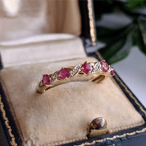 May include: A gold ring featuring seven oval-cut, vivid red gemstones and small diamonds. The ring has a twisted band design and is presented in a vintage blue jewellery box with a gold clasp. The ring is a beautiful piece of jewellery.