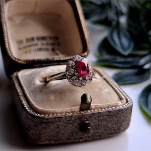 Op de afbeelding: Een vintage ring met een levendige rode edelsteen in het midden, omringd door kleinere heldere stenen, gezet in een zilveren band. De ring ligt in een open, antieke sieradendoos met een donkerbruine buitenkant en een beige binnenkant.