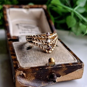 Könnte beinhalten: Ein goldener Ring mit drei gedrehten Bändern und drei kleinen Edelsteinen. Die Edelsteine sind ein klarer Diamant in der Mitte und zwei rosa Steine auf jeder Seite.