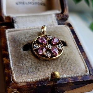 Può includere: Un ciondolo d'oro con pietre preziose rosa e bianche incastonate in un design circolare. Il ciondolo è in una scatola di gioielli in velluto marrone.