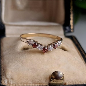 Puede incluir: Un anillo dorado con un diseño en forma de V, engastado con piedras preciosas claras y rojas alternas. El anillo se muestra dentro de un joyero vintage con interior beige y exterior negro.
