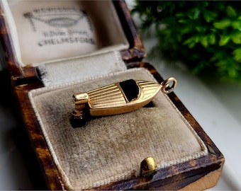 May include: A gold-colored boat charm with a black enamel detail, resting inside a brown velvet-lined box.