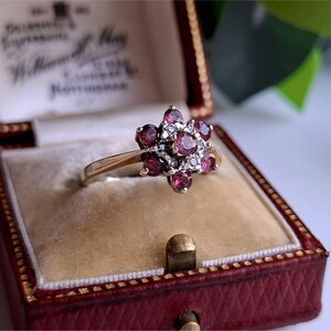 Può includere: Un anello d'oro con un design floreale, incastonato con pietre preziose rosse e piccole pietre bianche. L'anello è esposto in un portagioie rosso con chiusura dorata e interno color crema.