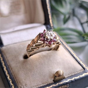 May include: A gold ring featuring a central cluster of small pink gemstones arranged in a floral pattern, with a single clear stone at the centre. The band is crafted with an engraved leaf motif along the sides, presenting a detailed botanical design.
