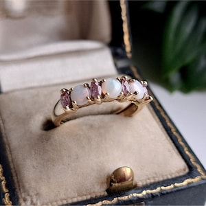 May include: A gold ring featuring three round white opals alternating with two marquise-cut pink gemstones. The stones are set in a prong setting along the top of the polished gold band, creating a delicate and symmetrical arrangement of colors.
