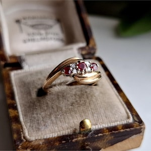 Könnte beinhalten: Ein goldener Ring mit drei roten Edelsteinen und kleinen weißen Steinen. Der Ring befindet sich in einer Vintage-Schmuckschatulle mit Schildpattmuster. Der Ring hat ein wirbelndes, elegantes Design.