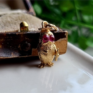 May include: A gold charm in the shape of a bird with ruby red eyes. The bird is holding a small scroll in its right wing.