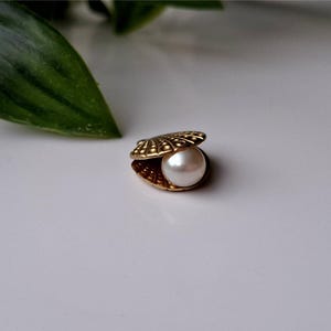 以下が含まれることがあります： 金色の貝殻の中に、大きくて丸い白い真珠が入っています。 貝殻には詳細な隆起とヒンジ付きの開口部があります。 背景はソフトホワイトで、左上隅に緑の葉があります。