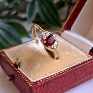 May include: A gold ring featuring a central oval red gemstone, accented by smaller white stones. The ring is presented in a red jewellery box with a cream interior, highlighting a vintage and sophisticated style.