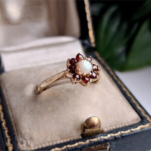 Könnte beinhalten: Ein Goldring mit einem zentralen, runden weißen Opal, der von acht tiefroten Granatsteinen in einem Blumenmuster umgeben ist. Der Ring ist schmal und poliert und zeigt ein klassisches Design mit einer Krappenfassung für jeden Edelstein.