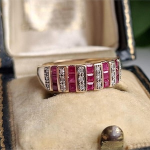 Könnte beinhalten: Ein goldener Ring mit einem rechteckigen Design mit abwechselnden Reihen von quadratisch geschliffenen roten Edelsteinen und kleinen, runden, klaren Edelsteinen. Der Ring wird in einer Vintage-Schmuckschatulle mit Samtinnenraum präsentiert.