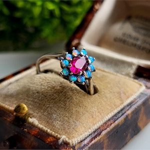 以下が含まれることがあります： 中央に楕円形のルビー、周囲に青いオパールを花のようにあしらったシルバーリング。リングは、ベルベットの内装が施されたヴィンテージのジュエリーボックスに収められています。