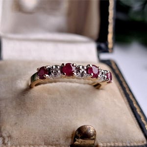 Peut inclure: Une bague en or avec trois rubis rouges ronds alternant avec deux petites pierres claires. La bague est montée sur un sertissage en métal, présentant un design multi-pierres avec une finition polie, présentée dans un écrin à bijoux doublé de velours bleu.